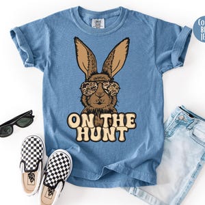 May include: Blue jean coloured t-shirt with a brown bunny wearing camouflage sunglasses. The text "ON THE HUNT" is printed below the bunny. The image also includes black sunglasses, checkered slip-on shoes, and blue jeans.
