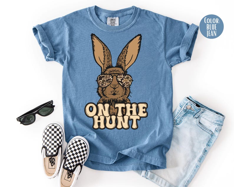 May include: Blue jean coloured t-shirt with a brown bunny wearing camouflage sunglasses. The text "ON THE HUNT" is printed below the bunny. The image also includes black sunglasses, checkered slip-on shoes, and blue jeans.