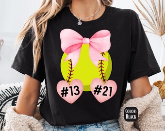 May include: Black t-shirt with a pink bow and a yellow softball with stitching. The softball has two pink hearts hanging from it with the numbers #13 and #21.