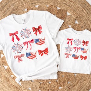 May include: Two white t-shirts with a red, white, and blue American flag design. The design includes fireworks, bows, and the outline of the United States.