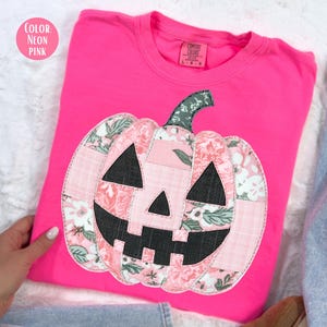 May include: Neon pink long-sleeve shirt with a patchwork pumpkin design. The pumpkin features floral and solid pink fabric with black accents for the eyes and mouth. The shirt is a vibrant color, perfect for fall.