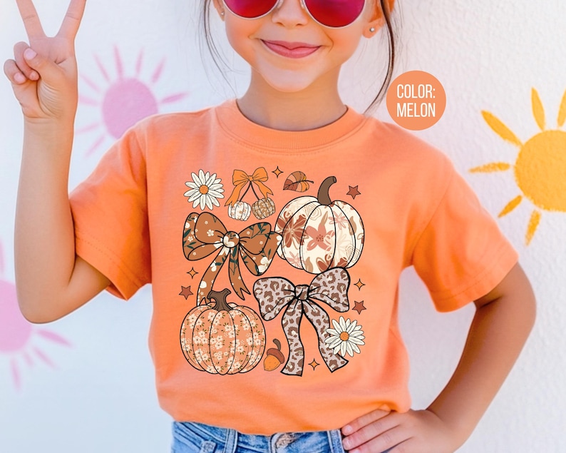 Peut inclure: T-shirt orange couleur melon avec un motif sur le th&egrave;me de l'automne. Le motif comprend des citrouilles, des n&oelig;uds, des fleurs et des feuilles dans des tons de marron, cr&egrave;me et orange.