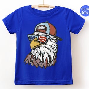 May include: Royal blue t-shirt featuring a graphic of an eagle wearing sunglasses with an American flag design. The eagle is also wearing a brown and blue baseball cap. The shirt is hanging on a wooden hanger.
