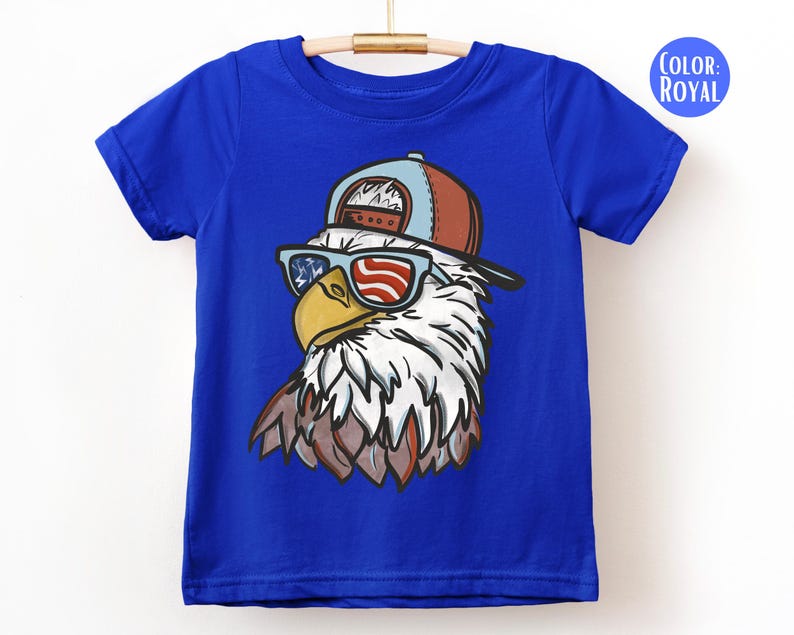 May include: Royal blue t-shirt featuring a graphic of an eagle wearing sunglasses with an American flag design. The eagle is also wearing a brown and blue baseball cap. The shirt is hanging on a wooden hanger.