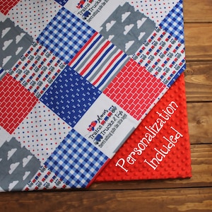 May include: A red, white, and blue patchwork baby blanket with a variety of patterns, including trucks, planes, and brick designs. The blanket is made of soft, comfortable fabric and is perfect for cuddling. The text "Trains, Trucks & Toys there's nothing quite like little boys!" is printed on the blanket. The blanket is paired with a red minky dot fabric.