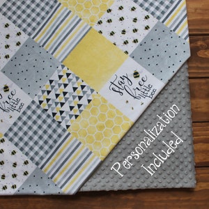 May include: A gray and yellow baby blanket with a patchwork design featuring bees, honeycomb, and geometric patterns. The blanket has the text "Stay free little bee" on one side and a gray minky fabric on the other side. The text "Personalization Included" is printed on the minky fabric.