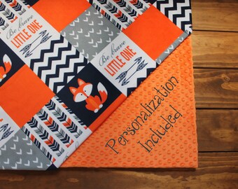 May include: A baby blanket with a patchwork design featuring orange, gray, and navy blue fabric. The blanket has a fox print, chevron pattern, and arrow designs. The text "Be brave little one" is printed on the blanket. The blanket is made of soft, plush fabric and is perfect for cuddling.  Personalization is included.