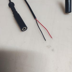 May include: A black audio cable with a 3.5mm female connector and exposed red and black wires. The cable is on a light-colored surface. The connector is cylindrical with a hole in the center.