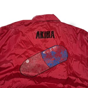 Peut inclure: Veste en nylon rouge avec un grand graphique de pilule bleu et rouge et le mot "AKIRA" imprim&eacute; au dos.