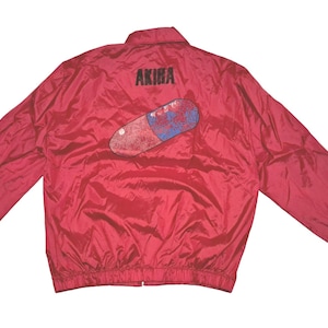 Peut inclure: Veste en nylon rouge avec une fermeture &eacute;clair noire et un graphique d'une pilule rouge et bleue avec le mot "AKIRA" imprim&eacute; au-dessus.