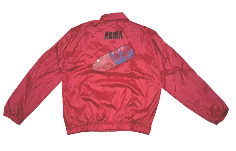 Peut inclure: Veste en nylon rouge avec une fermeture &eacute;clair noire et un graphique d'une pilule rouge et bleue avec le mot "AKIRA" imprim&eacute; au-dessus.