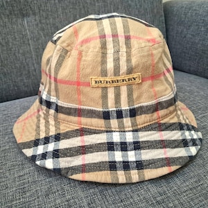 May include: A beige bucket hat with a classic plaid pattern in red, black, and white. The hat features a small rectangular label with the brand name "BURBERRY" in a dark font. The hat is a stylish accessory.