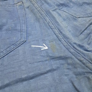May include: A close-up of a light blue fabric with a subtle checkered pattern. There is a small, light brown stain on the fabric.