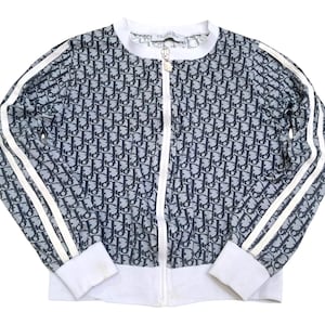 May include: A zip-up jacket with a repeating navy blue and gray pattern. The jacket has white stripes down the sleeves and white cuffs and collar. The zipper is white with a gold-colored pull.