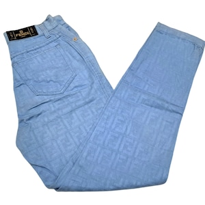 May include: Light blue jeans with a repeating pattern of the word "FENDI" in a darker blue color. The jeans have a black leather patch with the word "FENDI" in white lettering.