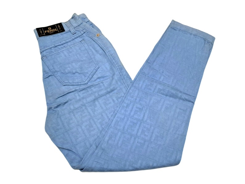 May include: Light blue jeans with a repeating pattern of the word "FENDI" in a darker blue color. The jeans have a black leather patch with the word "FENDI" in white lettering.