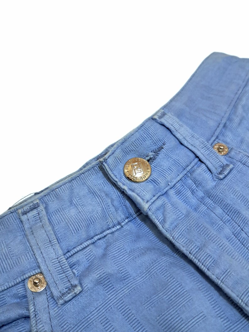 May include: Close-up of a pair of light blue jeans with a checkered pattern. The jeans have a button fly and a small metal button with the text "MADE IN USA" on it.