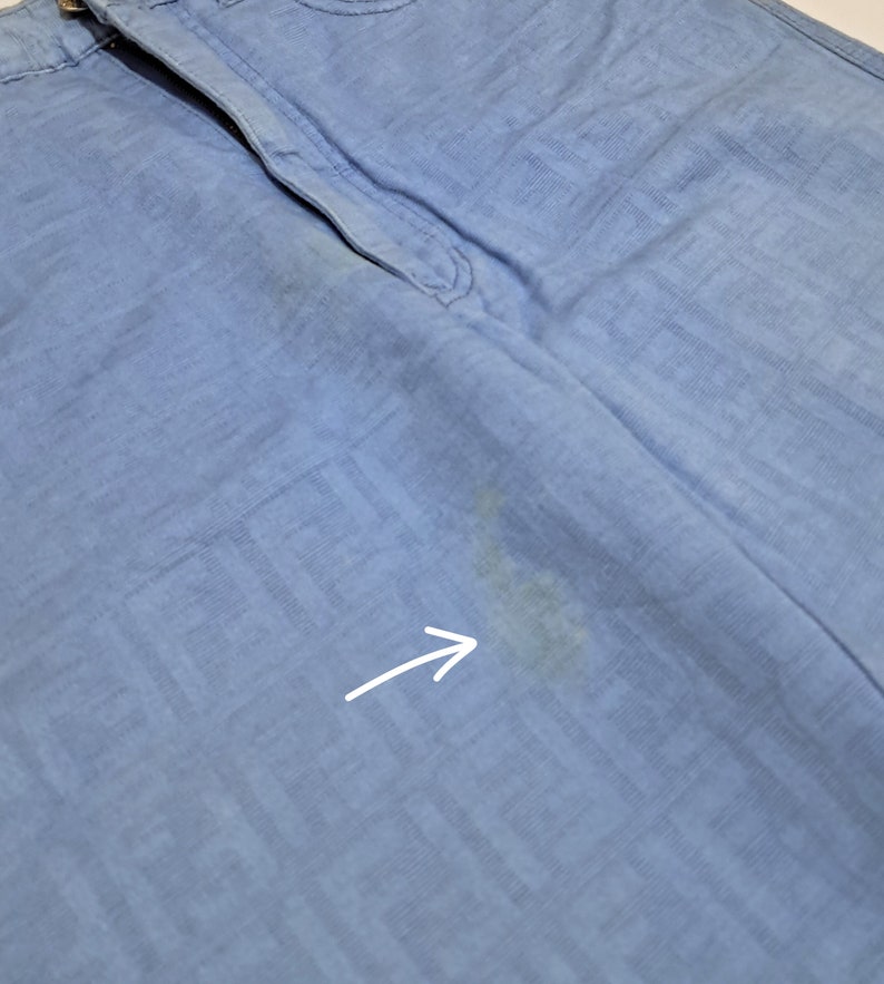 May include: Light blue pants with a subtle pattern and a small stain on the right leg. The stain is indicated by an arrow.