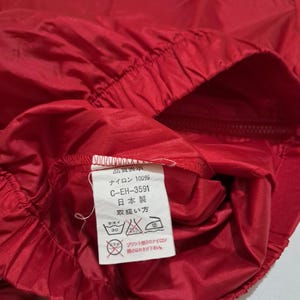 Peut inclure: Gros plan sur une veste en nylon rouge avec une &eacute;tiquette d'entretien blanche. L'&eacute;tiquette indique "Quality Display", "Nylon 100%", "C-EH-3591", "Fabriqu&eacute; au Japon" et "Instructions d'entretien".