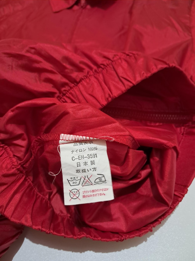 Peut inclure: Gros plan sur une veste en nylon rouge avec une &eacute;tiquette d'entretien blanche. L'&eacute;tiquette indique "Quality Display", "Nylon 100%", "C-EH-3591", "Fabriqu&eacute; au Japon" et "Instructions d'entretien".
