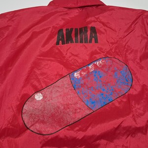 Peut inclure: Veste en nylon rouge avec un grand graphique de pilule rouge et le mot "AKIRA" imprim&eacute; en noir au dos.