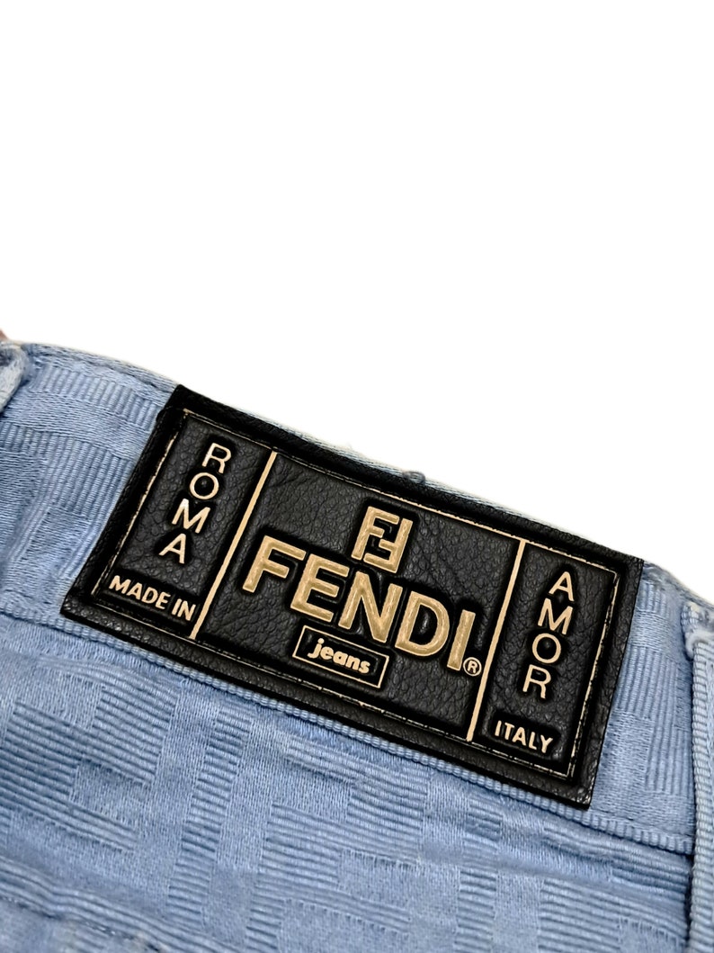 May include: A light blue pair of jeans with a black leather patch that says "FENDI Jeans" in gold lettering. The patch also has the words "Roma" and "Amor" in gold lettering. The patch also says "Made in Italy".