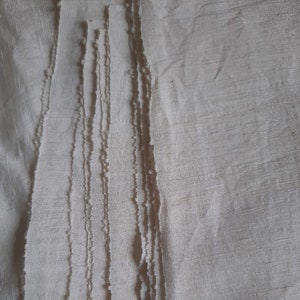Pu&ograve; includere: Primo piano di una pila di strisce di tessuto bianco con bordi sfilacciati. Il tessuto ha una texture sottile e un colore leggermente avorio.