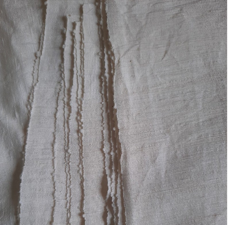 Pu&ograve; includere: Primo piano di una pila di strisce di tessuto bianco con bordi sfilacciati. Il tessuto ha una texture sottile e un colore leggermente avorio.