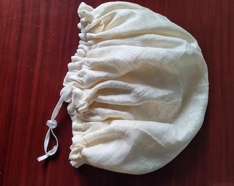 May include: Cream-colored drawstring bag made of textured fabric. The bag is gathered at the top with a white drawstring and a small, clear bead. The bag is likely used for storage or packaging.