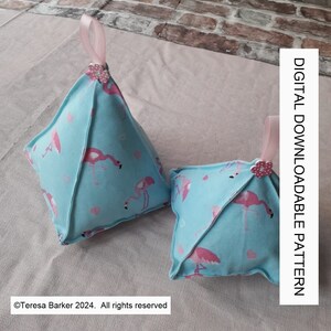 Puede incluir: Dos cojines de tela azul en forma de pirámide con estampado de flamencos. Los cojines tienen lazos de cinta rosa y adornos de flores rosas. El texto "DIGITAL DOWNLOADABLE PATTERN" está en el lado derecho de la imagen.
