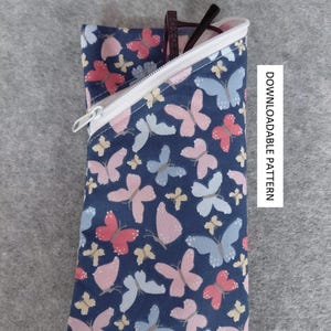 Könnte beinhalten: Ein marineblaues Brillenetui mit weißem Reißverschluss, gefüttert mit weißem Stoff. Das Etui ist mit einem Muster aus rosa, blauen und gelben Schmetterlingen verziert. Der Text "DOWNLOADABLE PATTERN" ist auf der rechten Seite sichtbar.