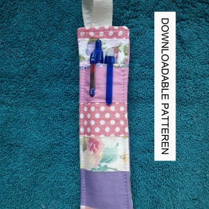 Könnte beinhalten: Ein rosa-weißer Stoff-Bleistifthalter mit einer Schlaufe zum Aufhängen. Der Halter hat ein Blumenmuster und Punkte. Er ist mit zwei blauen Stiften gefüllt.
