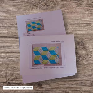 May include: Two white sheets of paper with sewing patterns for a quilted patchwork table runner. The top sheet shows a smaller version of the table runner with blue, green, and white geometric shapes. The bottom sheet has the text "PATTERN NUMBER #1056" and "SEWING PATTERN FOR A QUILTED PATCHWORK TABLE RUNNER."