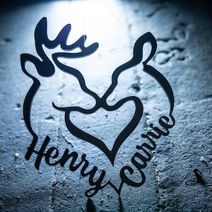 May include: A black silhouette of two deer heads facing each other with a heart in the middle. The names "Henry" and "Carrie" are written below the deer heads.