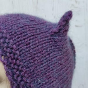 Puede incluir: Primer plano de un gorro morado tejido a mano con un detalle de oreja puntiaguda. El gorro está hecho de un hilo grueso y texturizado con sutiles reflejos azules. Un accesorio único y hecho a mano.