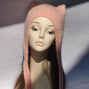 Könnte beinhalten: Eine hellrosa gestrickte Mütze mit Katzenohren. Die Mütze hat lange Bänder, die auf beiden Seiten herunterhängen. Die Mütze wird auf einem Schaufensterpuppenkopf mit langen braunen Haaren präsentiert.