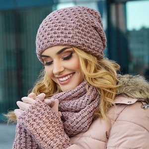 Op de afbeelding: Een bijpassende set gebreide winteraccessoires in een stoffige roze kleur. De set bevat een muts, een sjaal en vingerloze handschoenen. De persoon draagt een lichtroze gewatteerde jas met een met bont gevoerde capuchon.