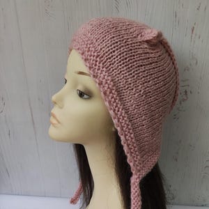 May include: A knitted pink hat with ear flaps and long ties. The hat is made of a chunky knit and is displayed on a mannequin head. The background is a light wood-paneled wall.