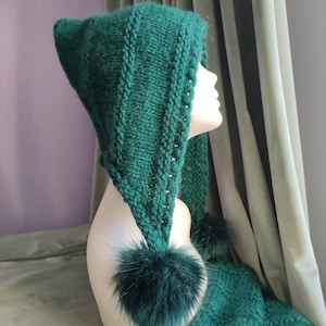 Peut inclure: Une cagoule verte tricotée avec une écharpe attachée et deux pompons duveteux. Le capuchon a un sommet pointu et est présenté sur une tête de mannequin. L'écharpe pend avec un pompon à l'extrémité. La texture tricotée est visible.