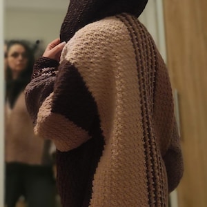May include: A hooded, oversized cardigan in shades of brown and beige. The back features vertical stripes and a textured knit pattern. The sleeves are long and the hood is a darker brown. The cardigan appears to be handmade.