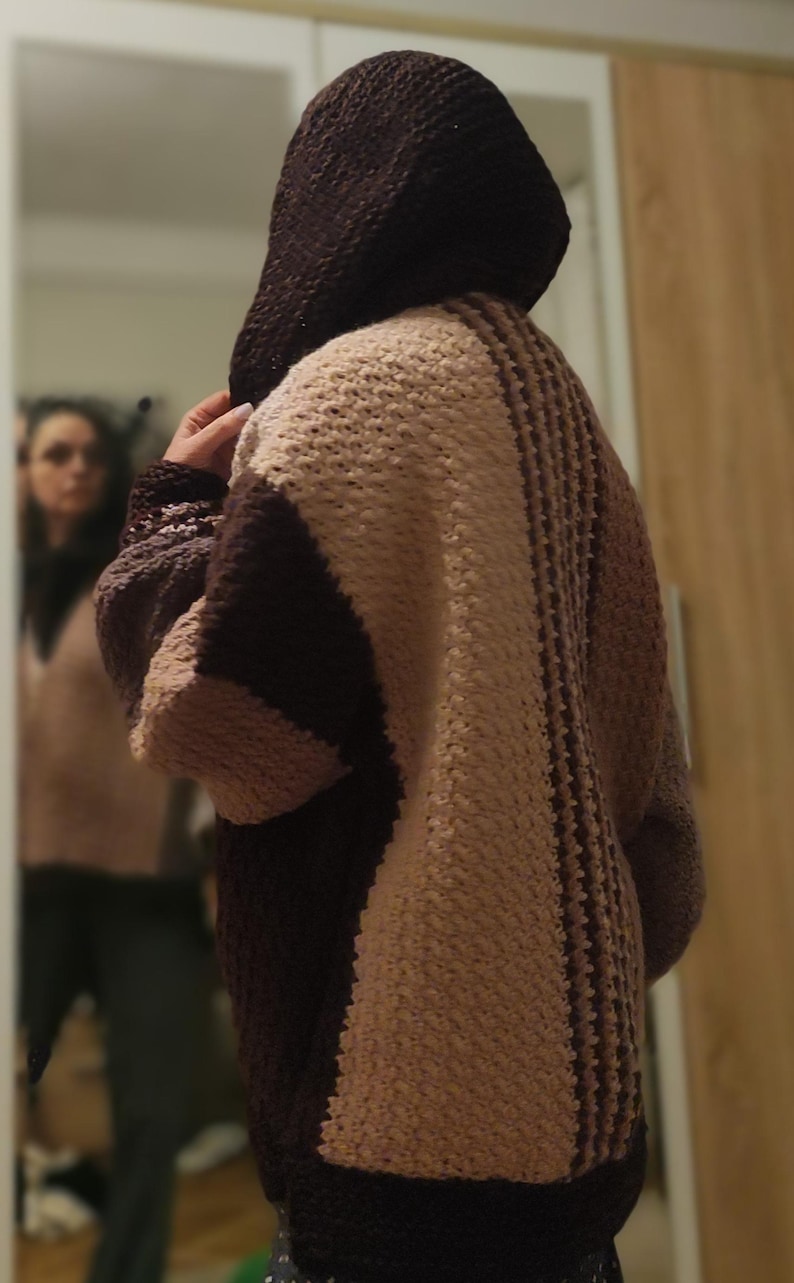 May include: A hooded, oversized cardigan in shades of brown and beige. The back features vertical stripes and a textured knit pattern. The sleeves are long and the hood is a darker brown. The cardigan appears to be handmade.