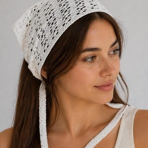 May include: A white, open-weave headscarf with long ties. The head covering is square-shaped and made of a delicate, crocheted material. The ties hang down the back. The headscarf is worn by a person with long brown hair.