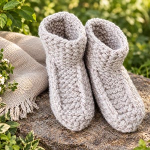 Könnte beinhalten: Ein Paar knöchelhohe Hausschuhe in hellem Beige mit grober Strickstruktur. Die Hausschuhe verfügen über eine abgerundete Zehenpartie und einen dicken, gerippten Bündchenabschluss, was ein gleichmäßiges, handgestricktes Erscheinungsbild vermittelt.