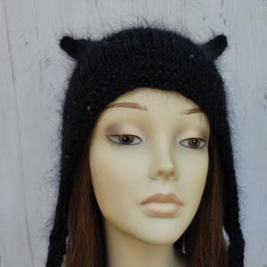 Peut inclure: Un bonnet noir en tricot avec de petites protubérances en forme d'oreilles. Le chapeau a de longues attaches tressées et est orné de petits sequins scintillants. Le chapeau est présenté sur une tête de mannequin avec des cheveux bruns.