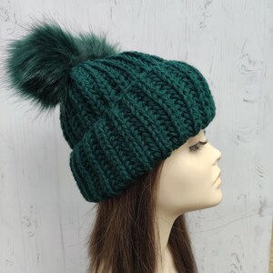 以下が含まれることがあります： 大きなフェイクファーのポンポンが付いた、濃い緑色のニット帽。帽子は厚手のリブ編み素材でできており、折り返しのつばが付いています。ポンポンは同じ緑色です。スタイリッシュな冬のアクセサリーです。