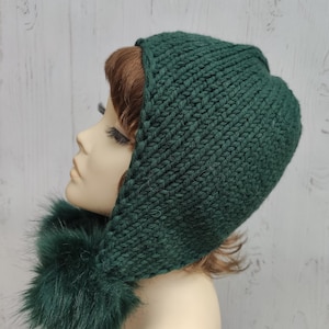May include: A dark green knitted hood with a faux fur trim. The hood is made of a thick, textured yarn. The faux fur is a matching dark green colour, giving a warm and cosy appearance.