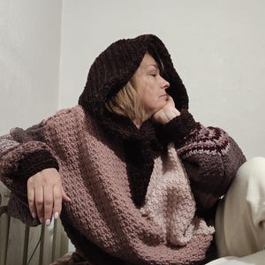 May include: A cozy, oversized hooded sweater in shades of brown and mauve. The sweater features a large hood, wide sleeves with patterned cuffs, and a textured knit design. The person is wearing the sweater.