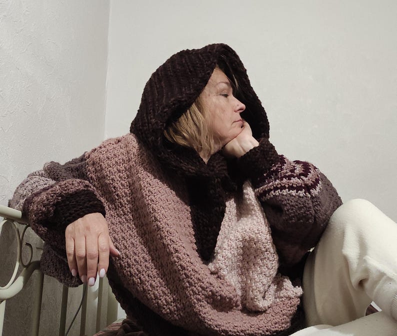 May include: A cozy, oversized hooded sweater in shades of brown and mauve. The sweater features a large hood, wide sleeves with patterned cuffs, and a textured knit design. The person is wearing the sweater.