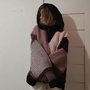 May include: A hooded, oversized crochet sweater in shades of brown, pink, and lavender. The sweater features a color-blocked design with a large hood and a relaxed fit. The sleeves are long, and the garment appears to be made of a chunky yarn.