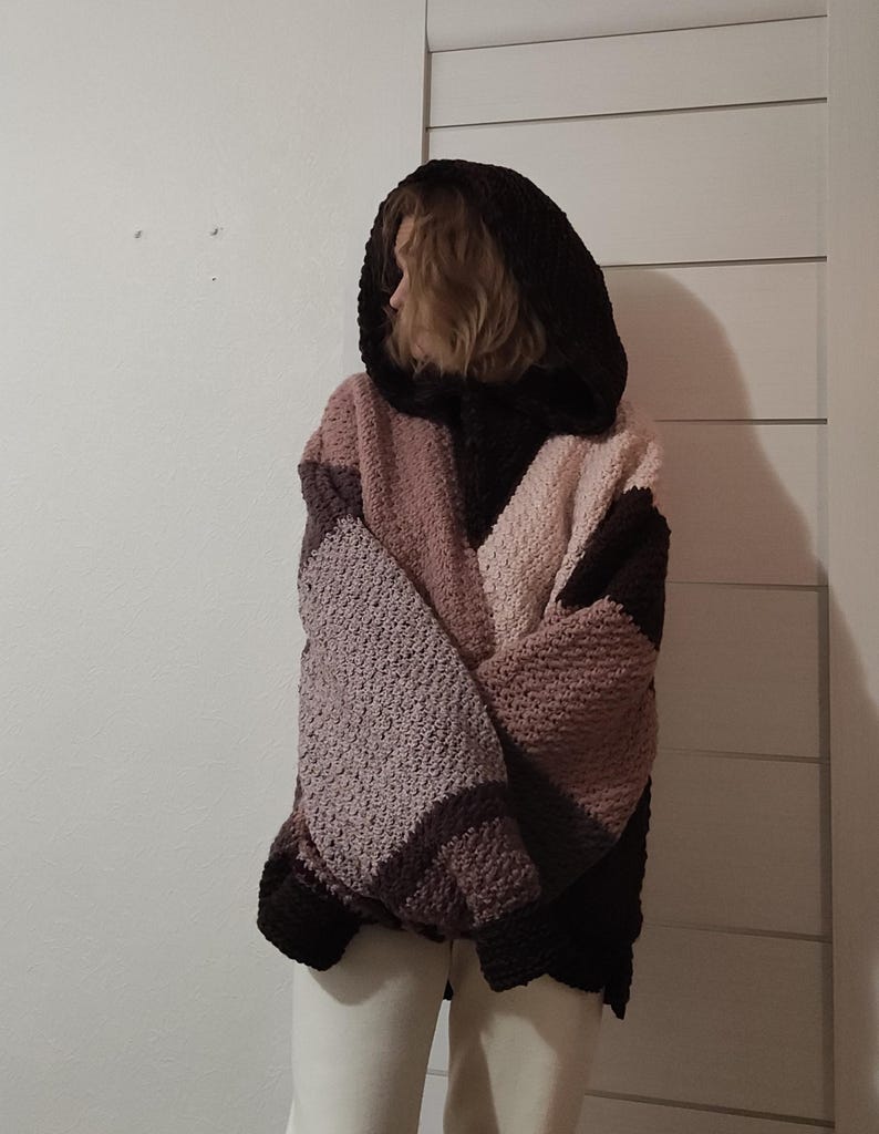 May include: A hooded, oversized crochet sweater in shades of brown, pink, and lavender. The sweater features a color-blocked design with a large hood and a relaxed fit. The sleeves are long, and the garment appears to be made of a chunky yarn.